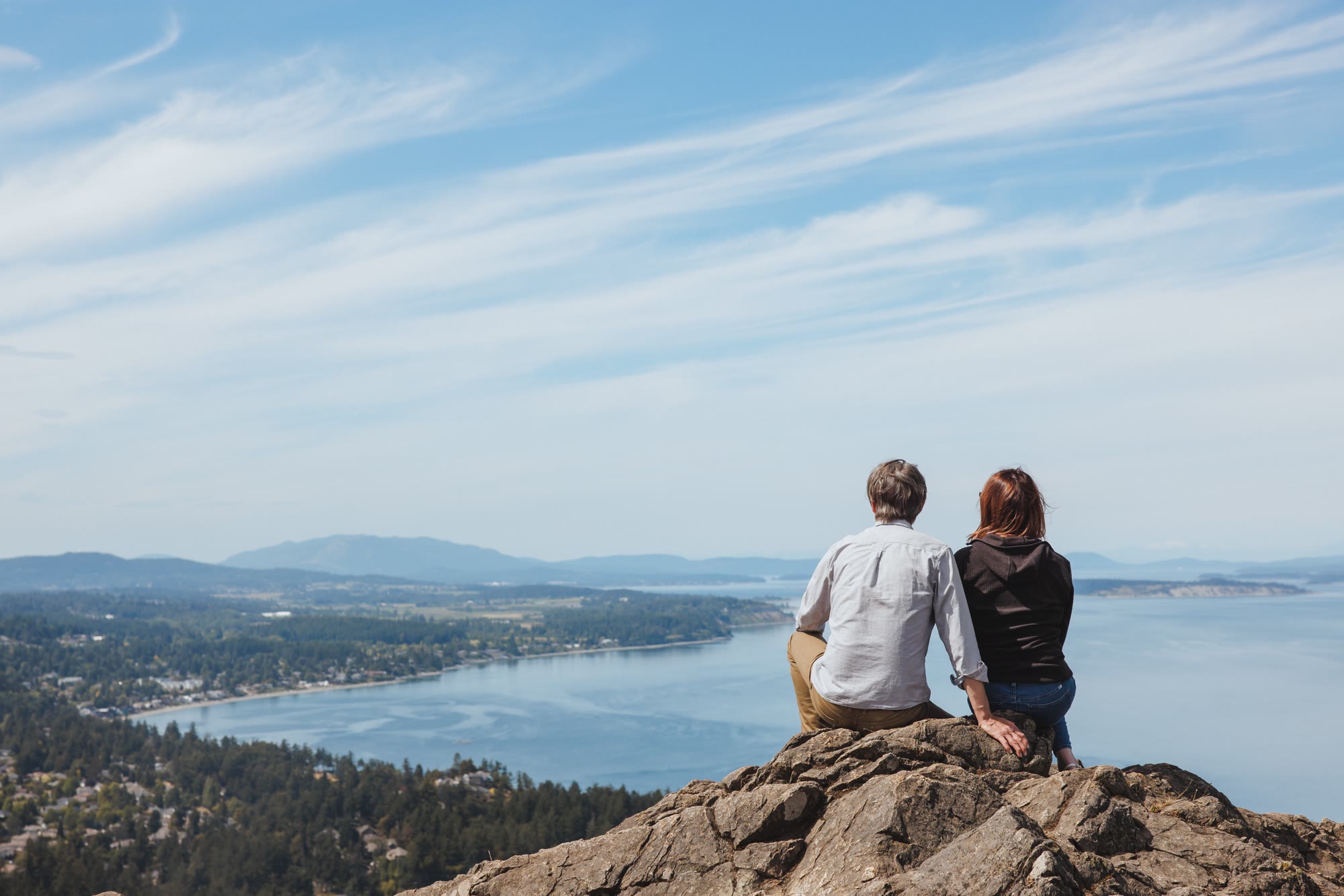 Two people sitting on the rocks at the summit of Mount Douglas, looking out over Cordova Bay and the islands