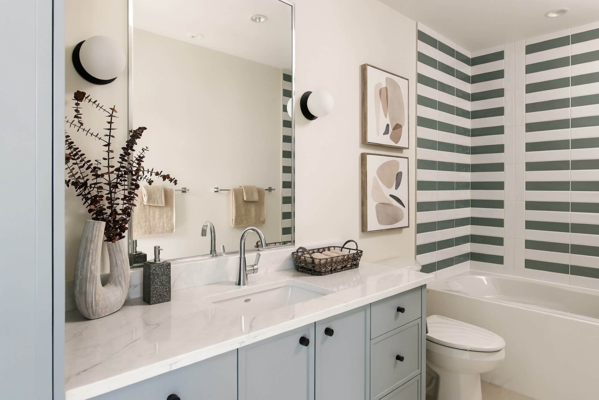 A bathroom with sage and white striped tile, a pale blue vanity and a quartz counter