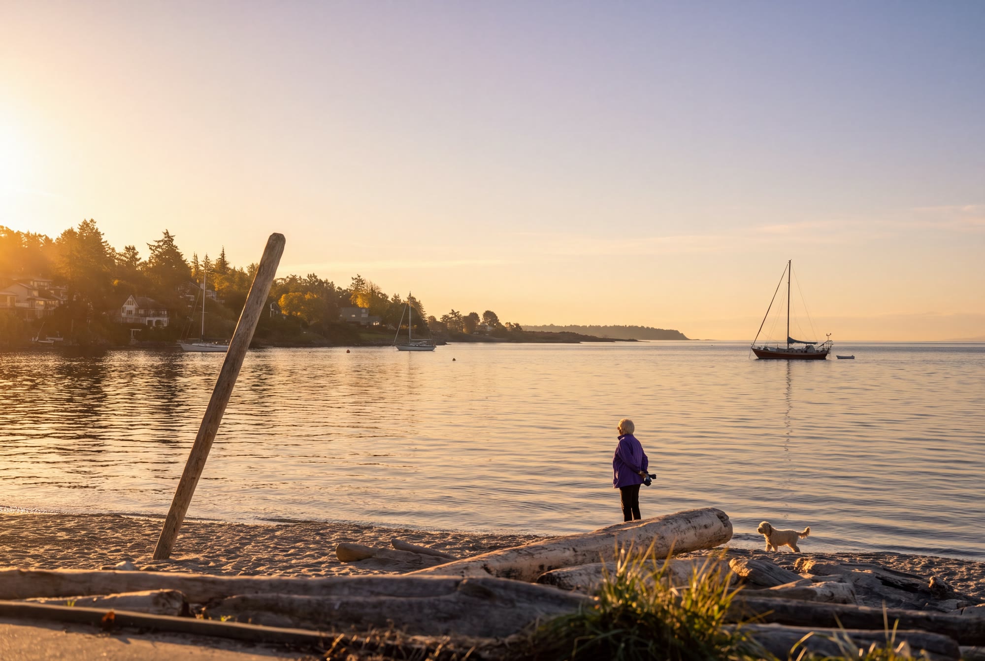 A woman and her dog on the sand at Cadboro Bay at golden hour, looking out at a moored sailboat